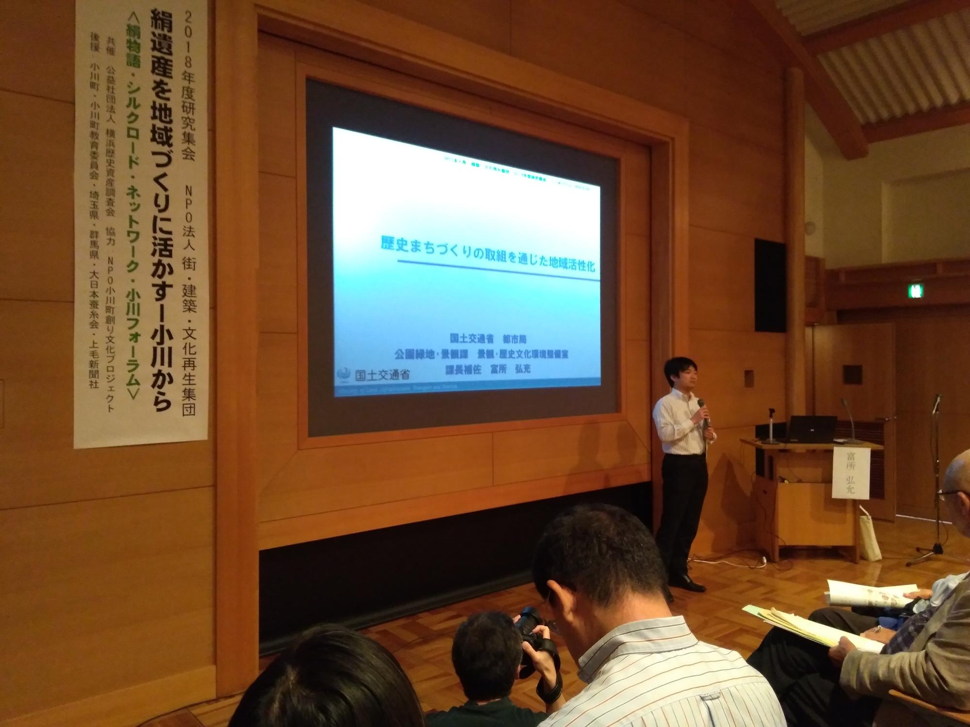 前方のスクリーンの前に男性がマイクを持ちたっている写真