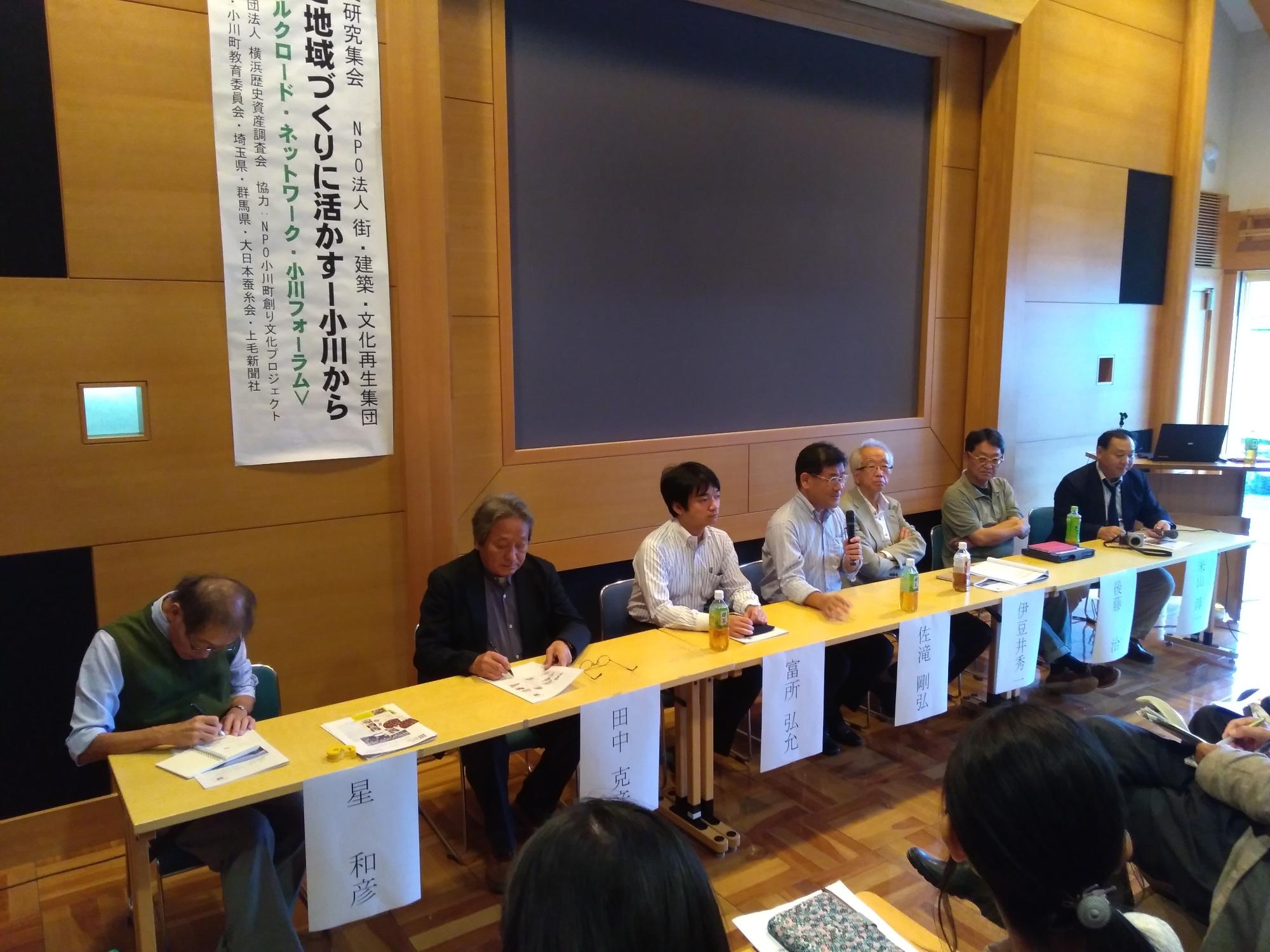 席次が書かれた長机に7名の人が座り真ん中の男性がマイクを持ち話をしている写真