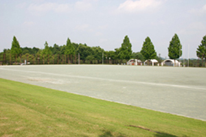 遠くに緑色に染まった木々が見える芝生とクレイ舗装された小川町総合運動場の写真