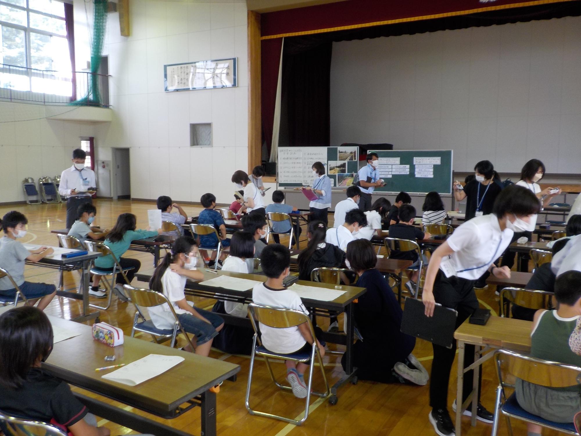 体育館で小学生たちが長机の前の椅子に座って防災授業を受けている写真