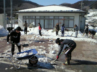 家の前にできた雪の山を女子学生が竹ぼうきを持ち、男子学生はスコップで一輪車に雪を載せて作業をしている様子を撮影した写真