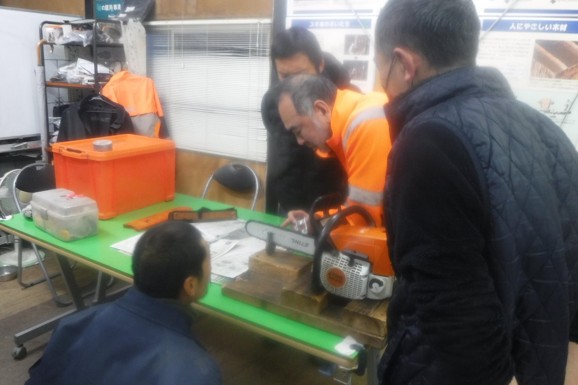 台の上に置かれた小型のチェーンソーを見ながら講習を受けている人々の写真