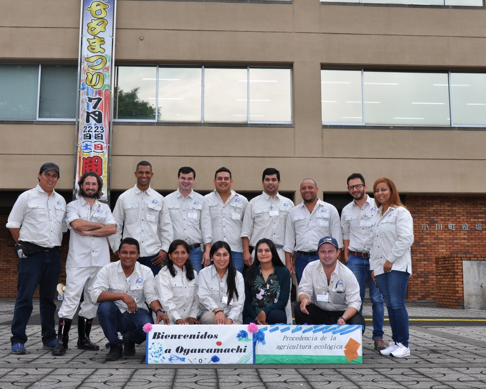 小川町役場を背景に女性3名を挟んで男性2名が前列に座り、男性8名と女性が後列に並んで母国語で書かれたプレートを持ち記念撮影をしている写真