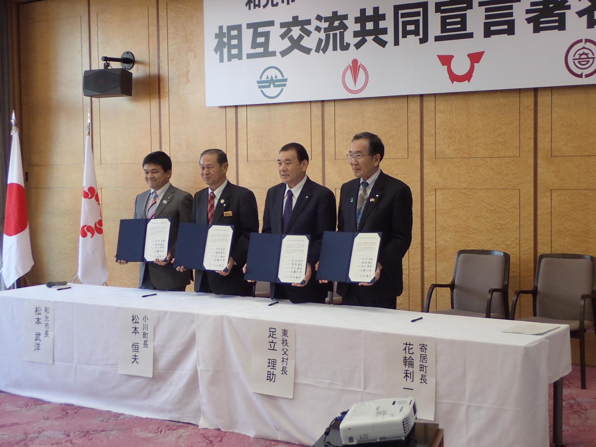 壁に「和光市と3町村（小川町・東秩父村・寄居町）との相互交流共同宣言署名式」と書かれた幕の前で4人の市町村長が協定書を持って並んでいる様子をななめ前から撮影した写真