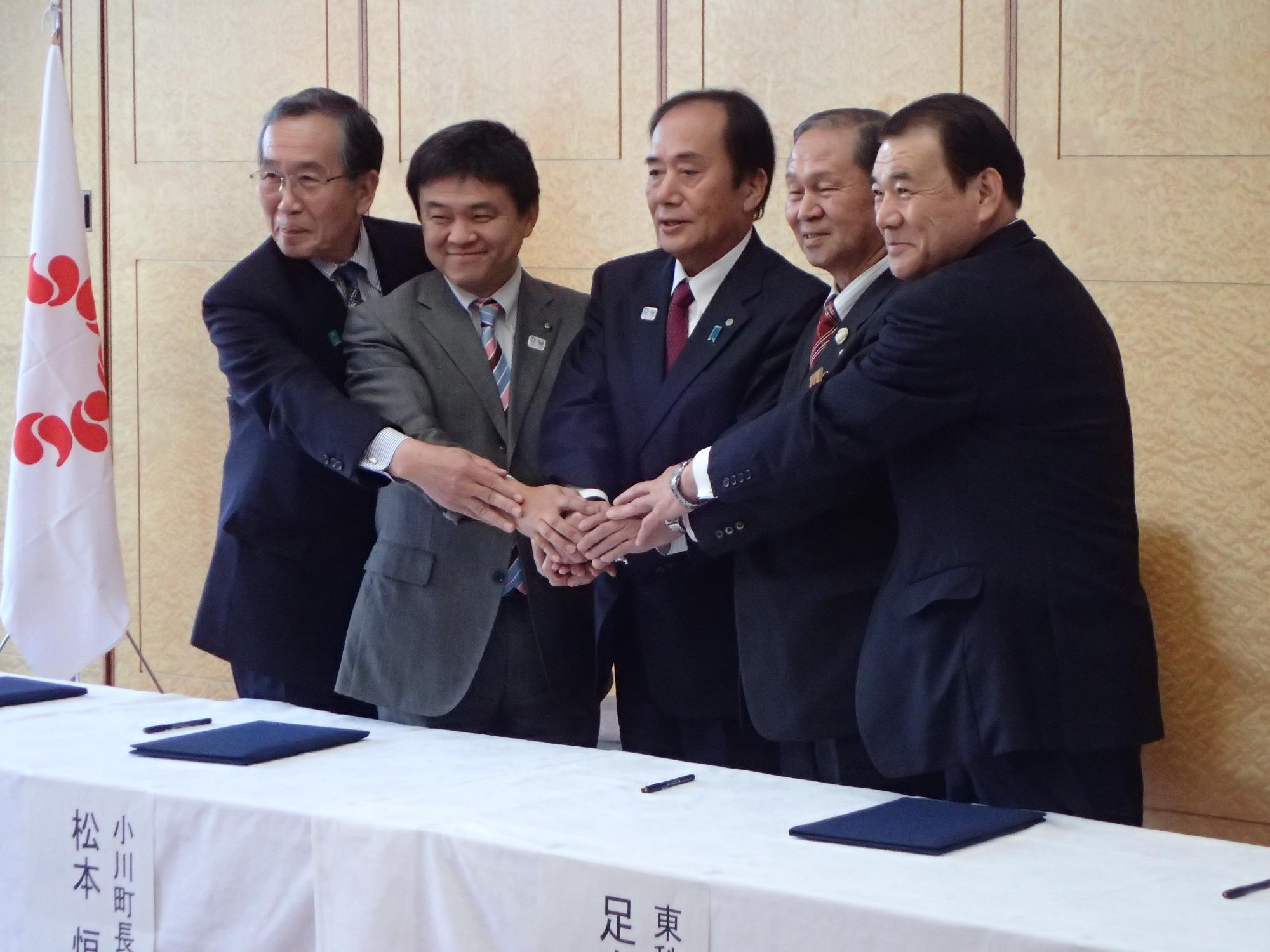 4人の市町村長の間に立つスーツ姿の男性を中心に5人で手を添えて正面を見て微笑んでいる様子を撮影した写真
