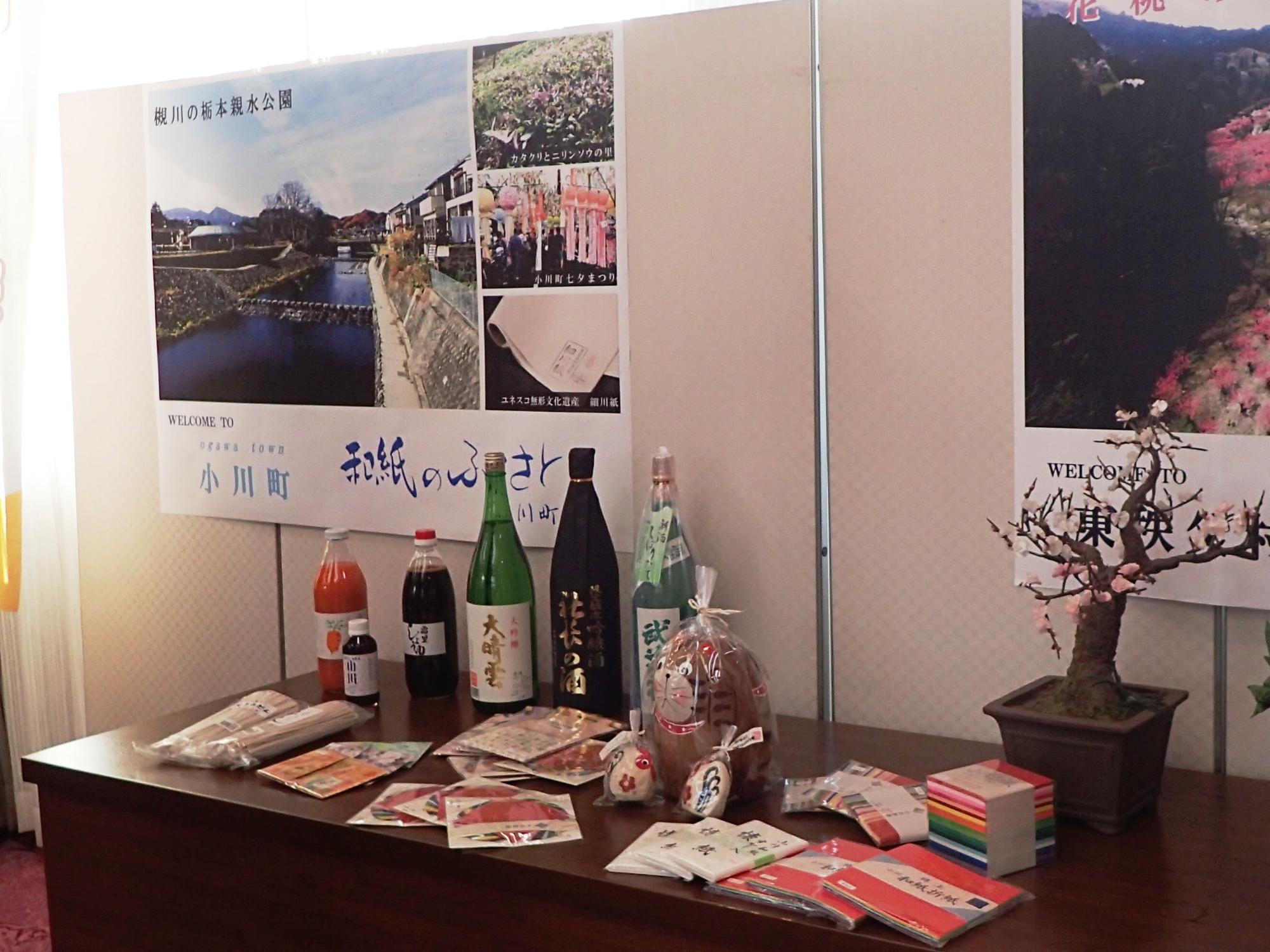 白いパネルに小川町、東秩父村のポスターが貼られていてその前に設置されたテーブルにはお酒やおしょうゆ、折り紙他、3町村の特産品が並べられている様子を撮影した写真