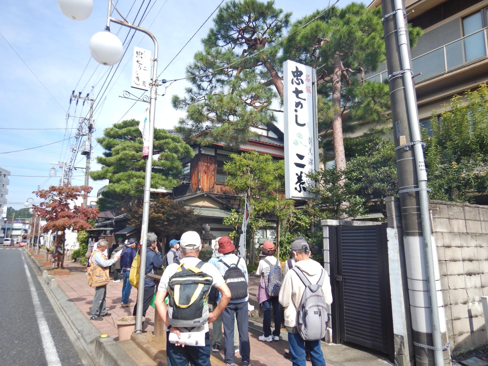 立派な松の木が生えている趣のある家の前で立ち止まって見学をしている参加者達の写真