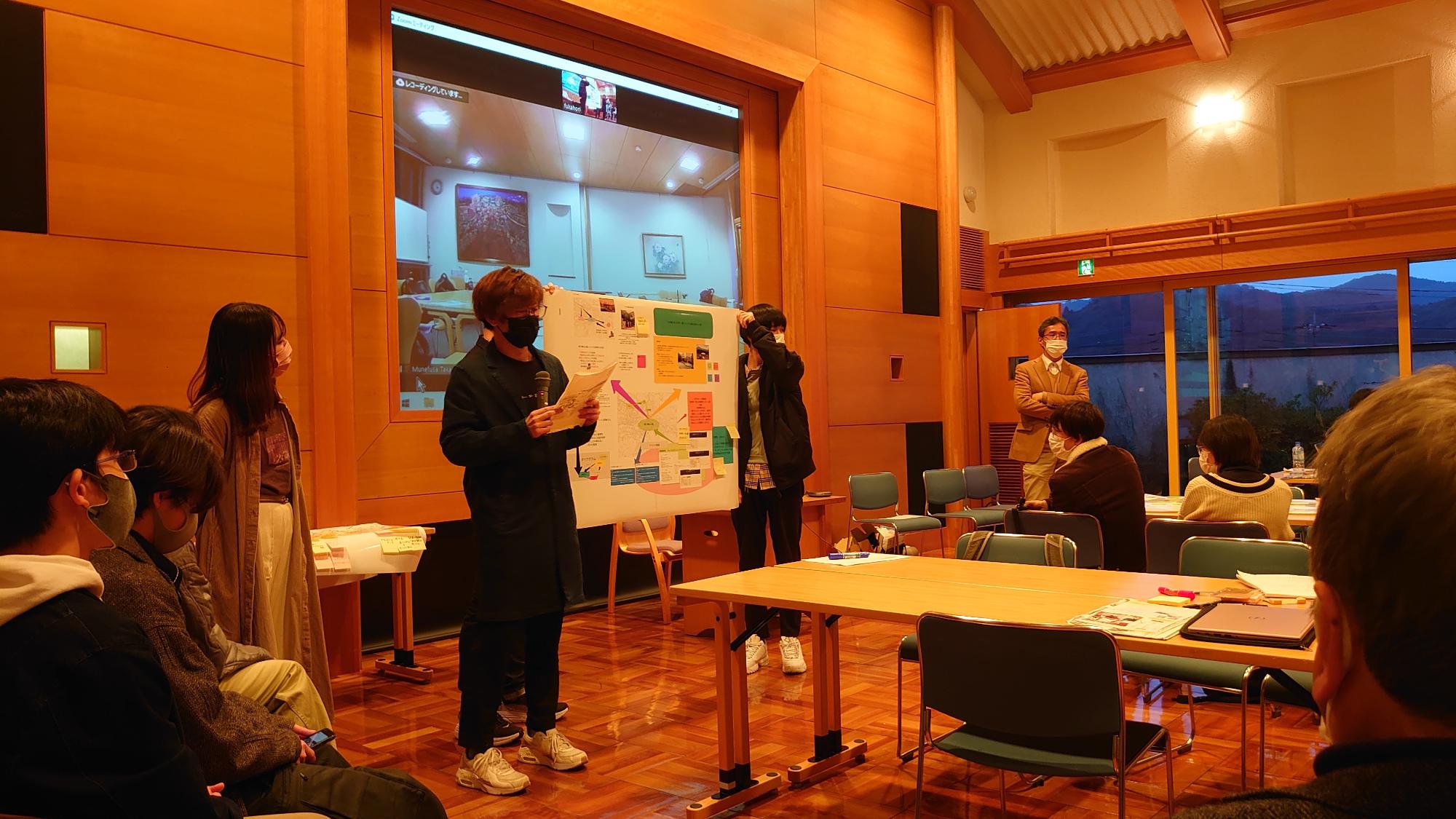 前方のホワイトボードを使って男性が説明をし、関係者2名が両脇にたっている発表会の写真