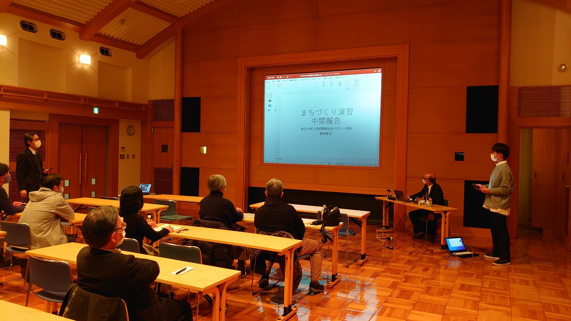 前方のスクリーンが写し出され説明を聞いている参加者と意見を述べているグレーの上着を着た男性の写真