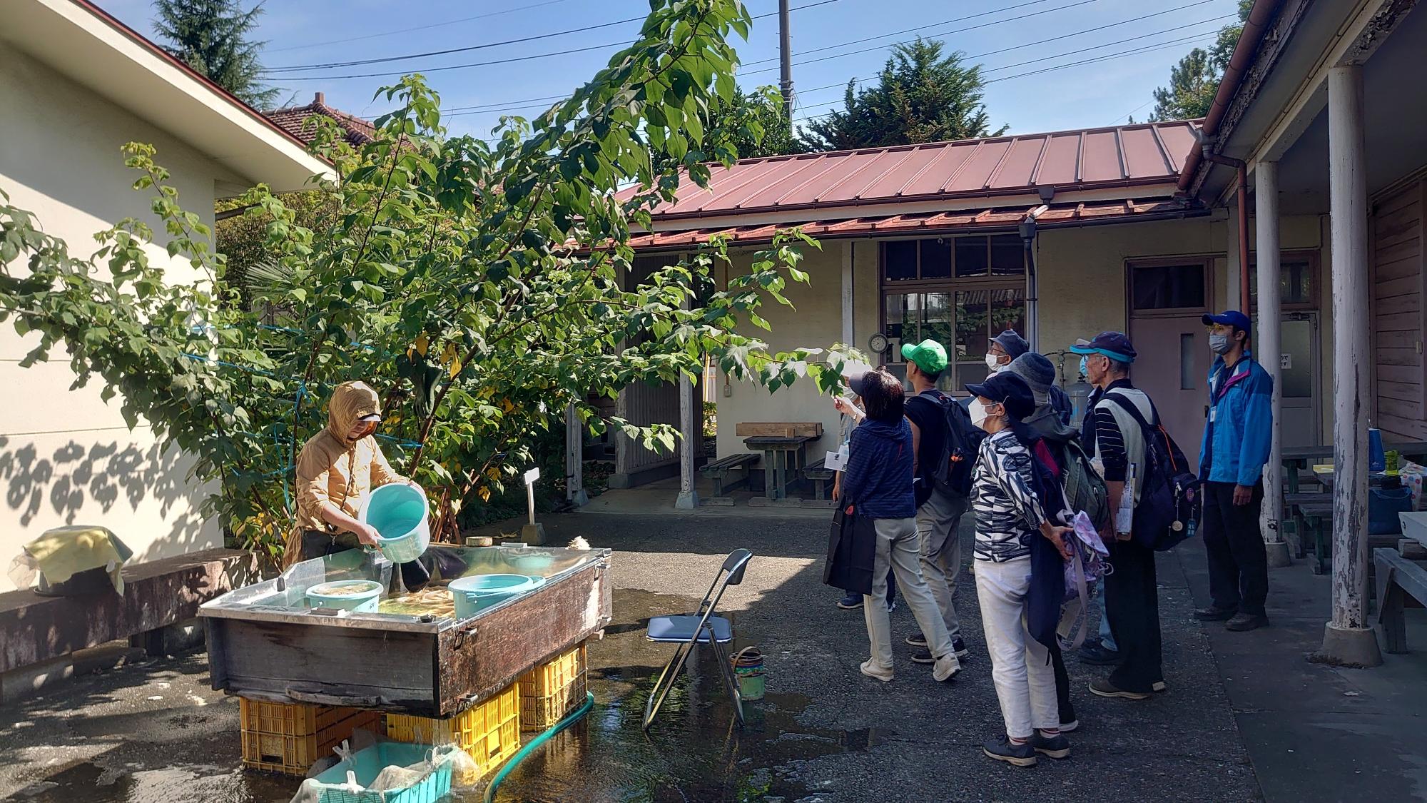 屋外でバケツを使って水作業をしている方がいる場所を見学している参加者達の写真