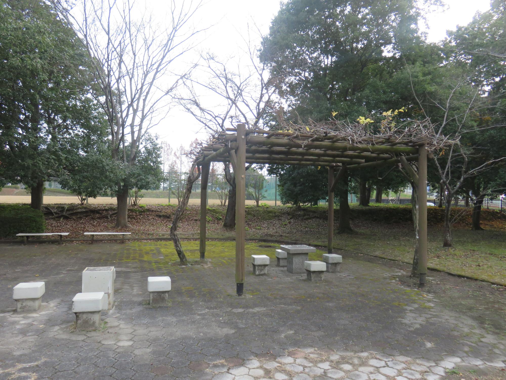 周りに木々が植えられ、奥にベンチ、手前に石で出来た椅子とテーブル、木製の藤棚が設置されている休憩スペースの写真