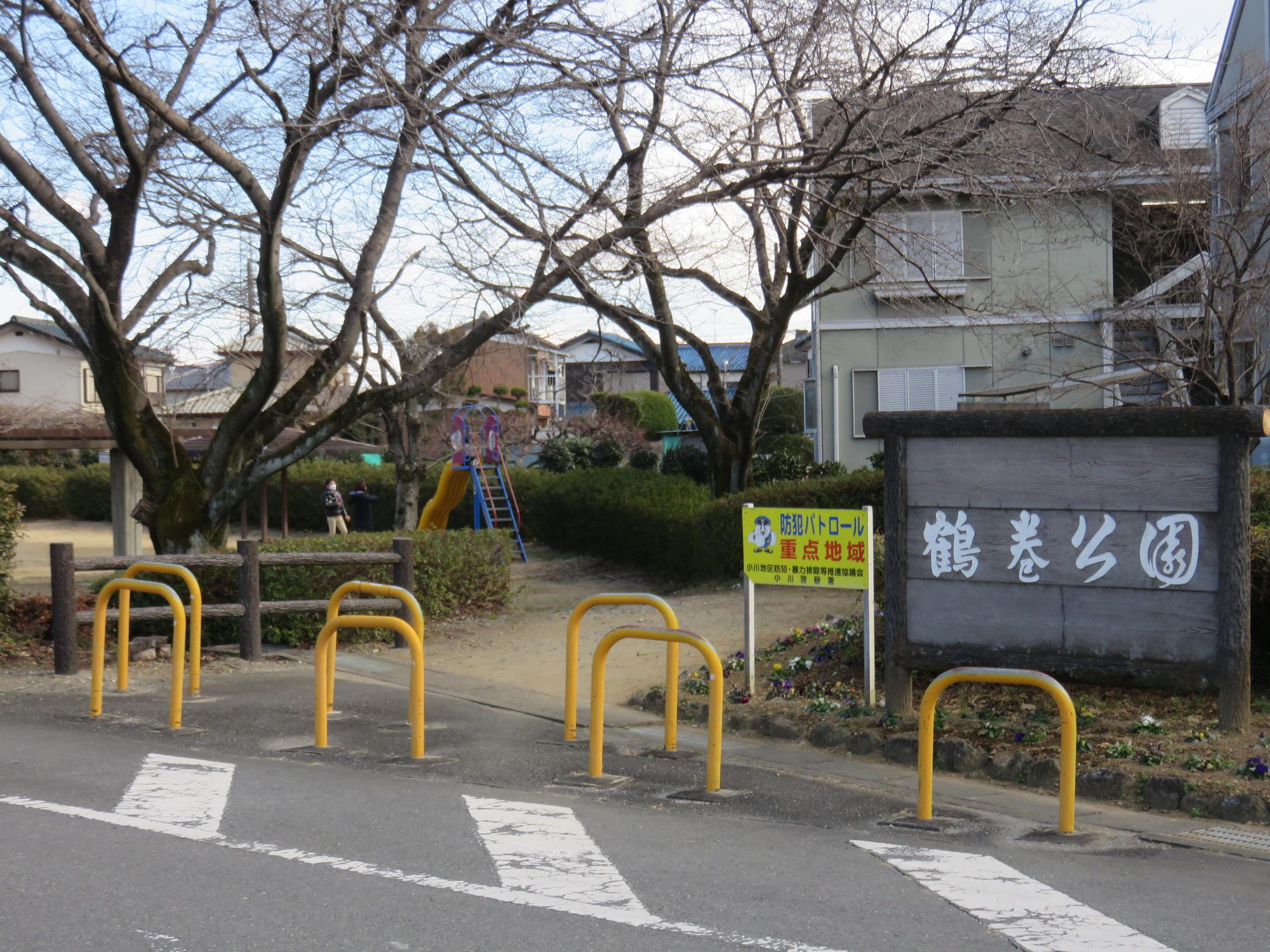 奥に滑り台が見える公園入口に車両進入防止柵があり、白い字で「鶴巻公園」と書かれた園名版が設置されている公園入口付近の写真