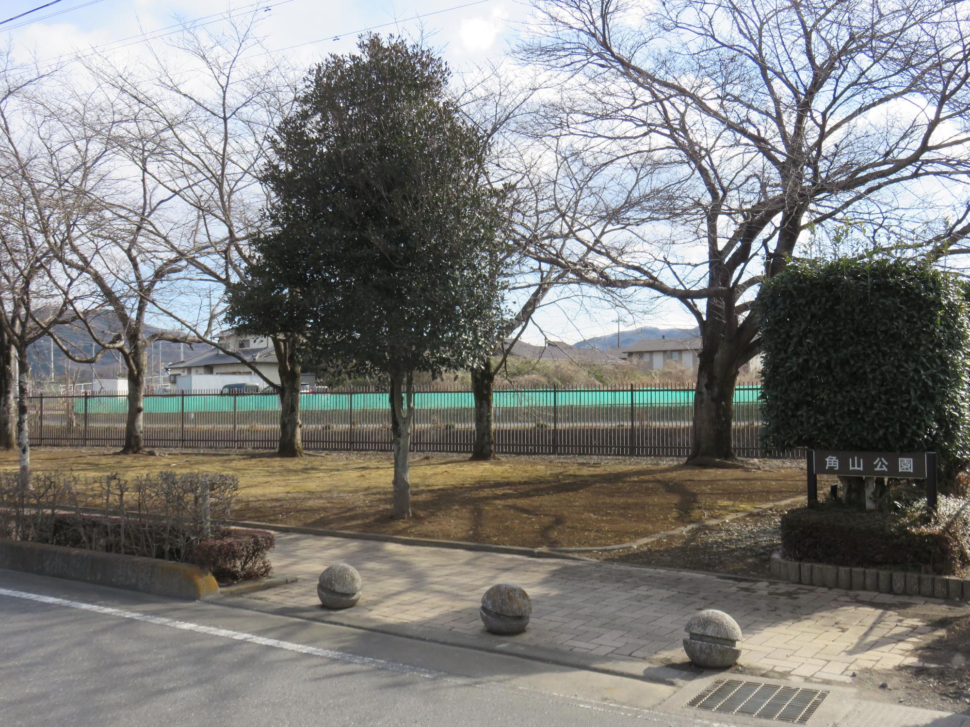 道路沿いの黒色のフェンスに囲まれた敷地内に間隔をあけて大きな木が立ち並んでいる角山公園の写真