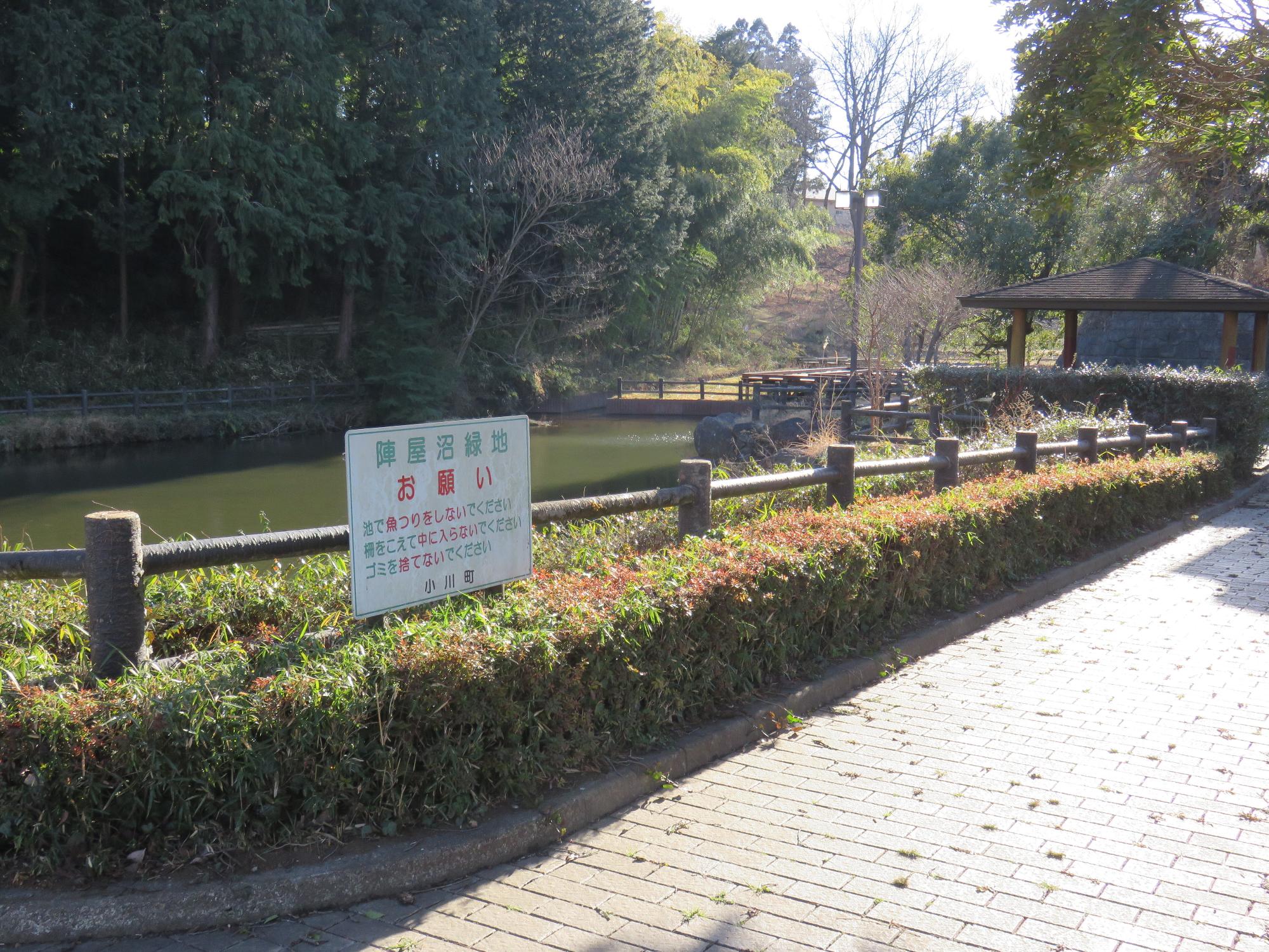 遊歩道や生垣に木の柵で囲まれた池の奥に東屋が設置された陣屋沼緑地の写真