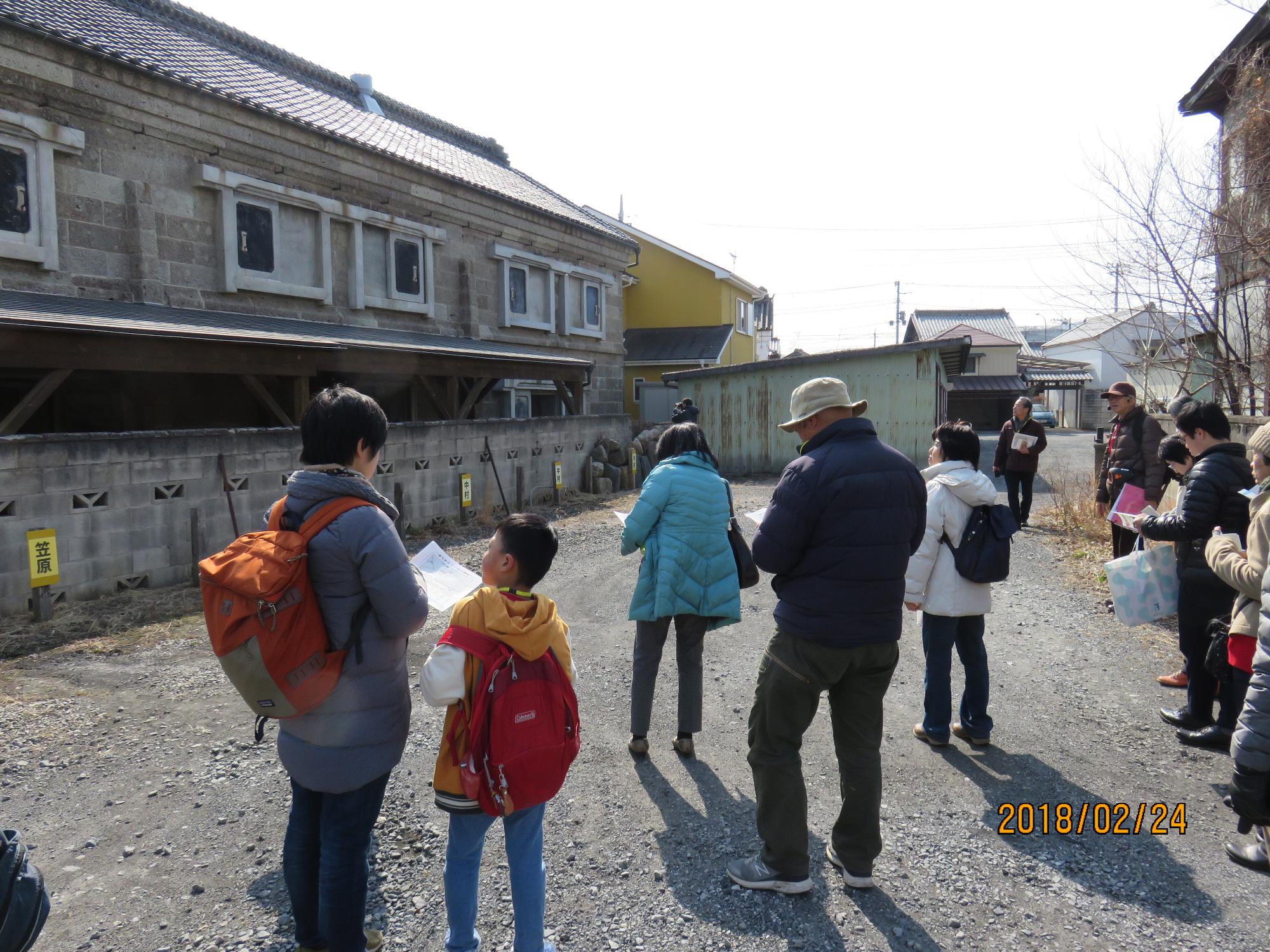 ダウンジャケットを着た参加者達が集まり、左側に建っている大きな蔵のような建物を見学している様子の写真