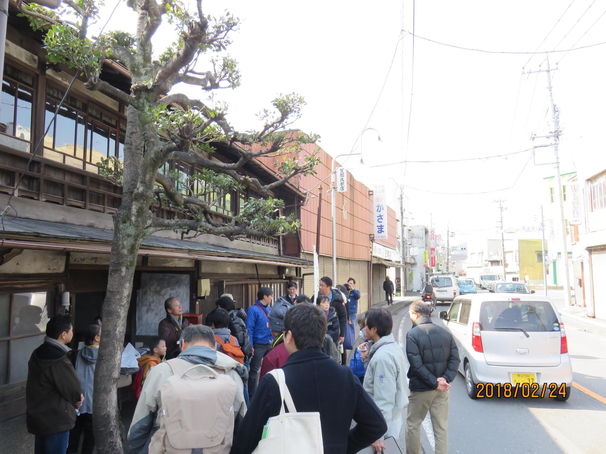 交通のある道路に面して建っている趣のある家の前に参加者達が集まっている写真