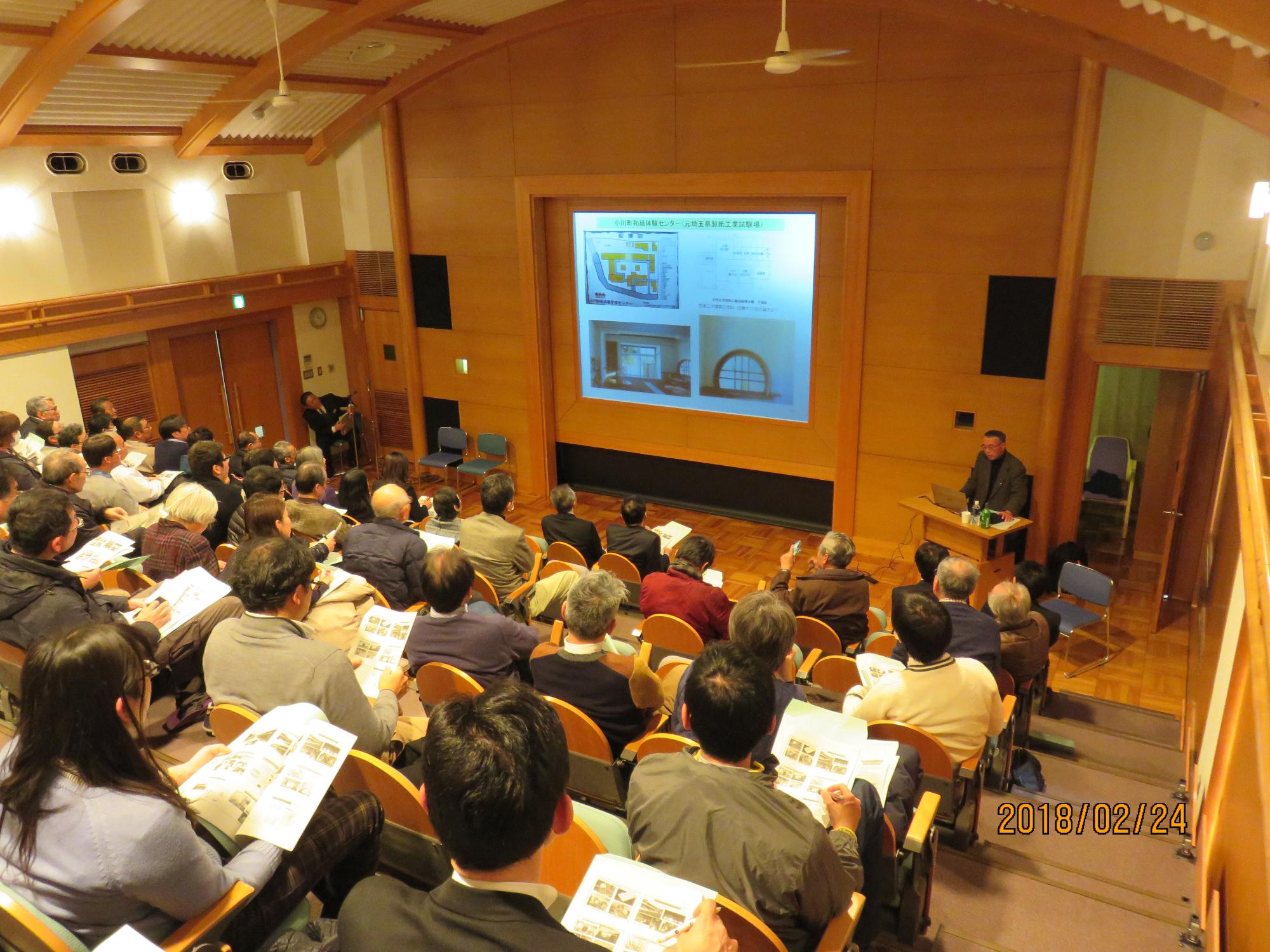 観客席が階段状になっている広いホールの前方に資料が映し出され、演台で講演を行っている苅谷氏の話を参加者達が席に座り聞いている様子の写真