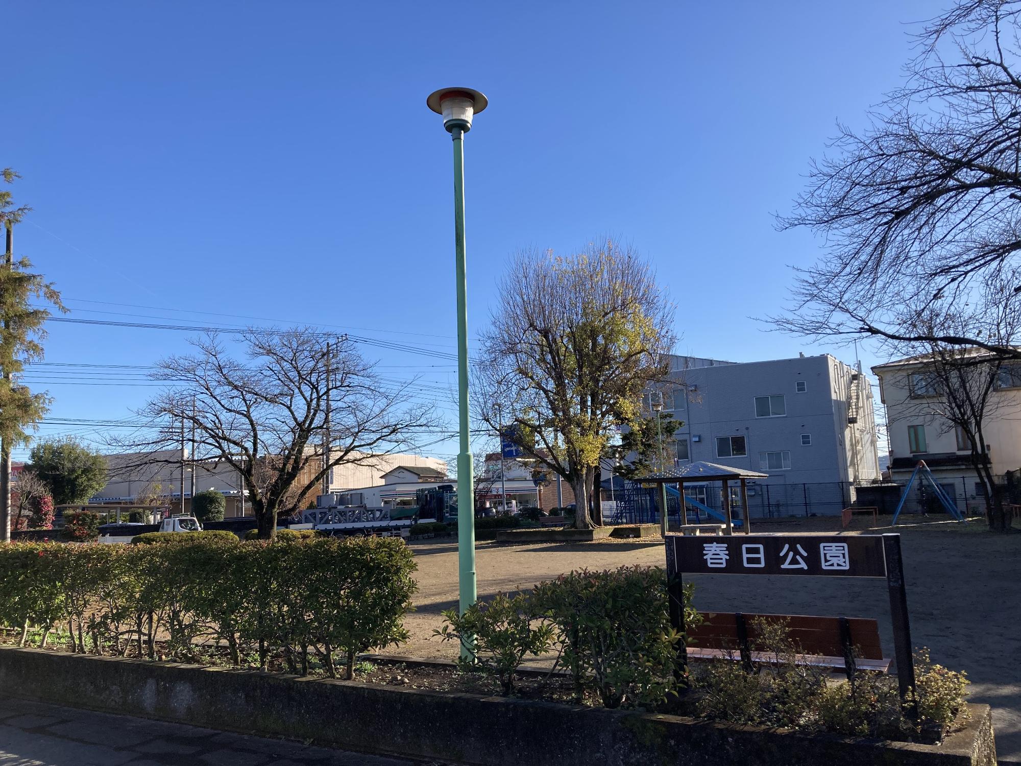 コンビニや住宅街の中にあり、間隔をあけて樹木が植えられた公園内に東屋やブランコ、複合遊具などが設置された春日公園の写真