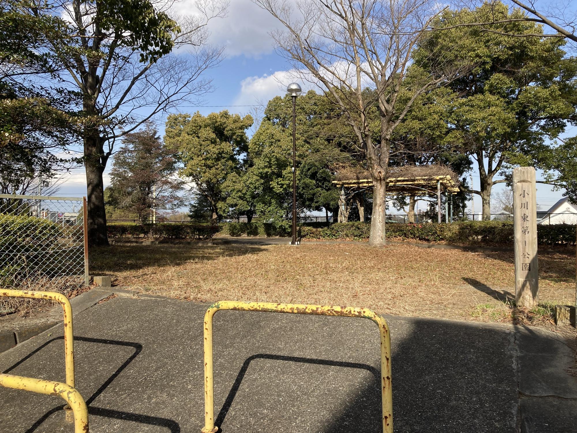 縦書きで「小川東第1公園」と書かれた木製看板が設置された奥に、木々に囲まれ東屋が設置された公園内の写真