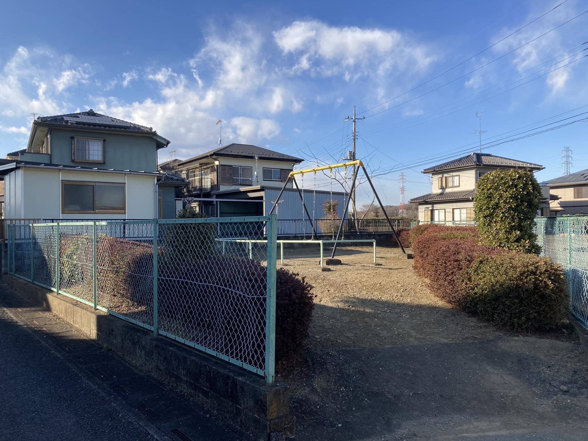 住宅街に隣接した緑色のフェンスと生垣に囲まれた敷地内にブランコが設置された中爪東公園の写真