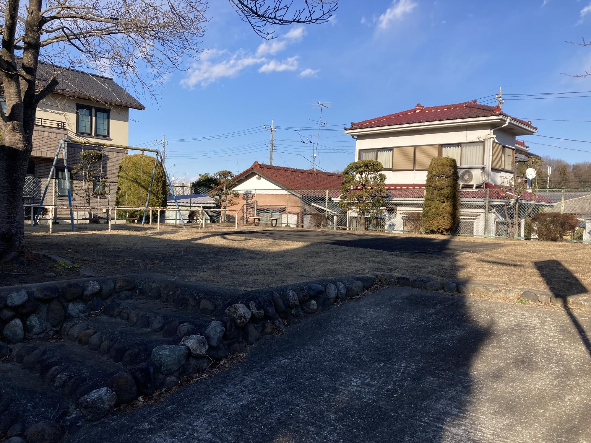 住宅街に隣接しフェンスに囲まれた敷地内にベンチやブランコが設置された梶山公園の写真