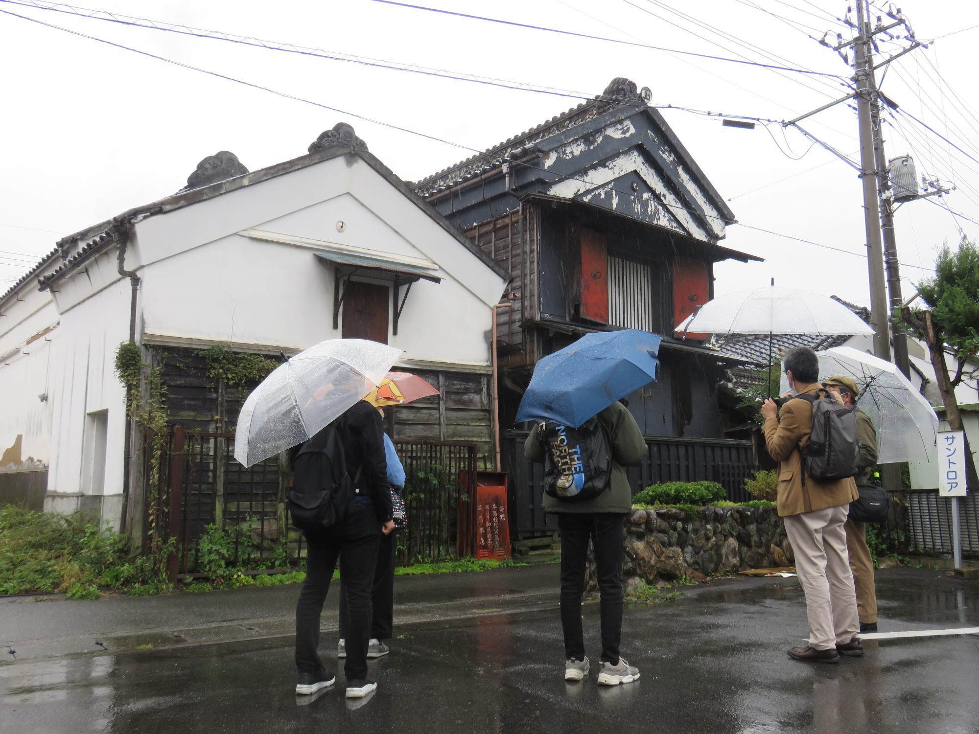 5名の方が傘をさしながら建物の前で説明を聞いているまち歩きの写真
