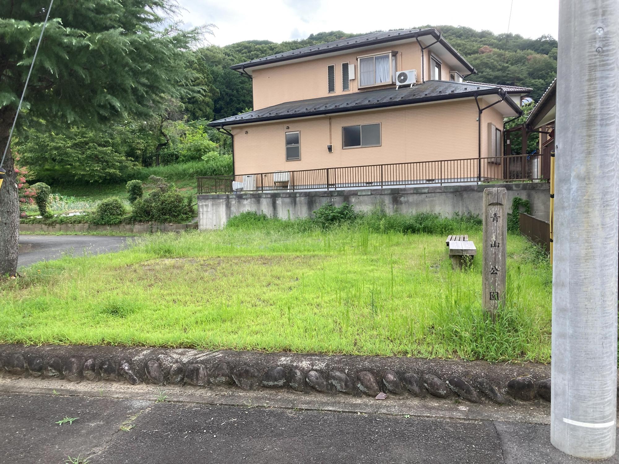 民家に隣接した一角にある草が生えた敷地内に2台のベンチが設置された青山公園の写真