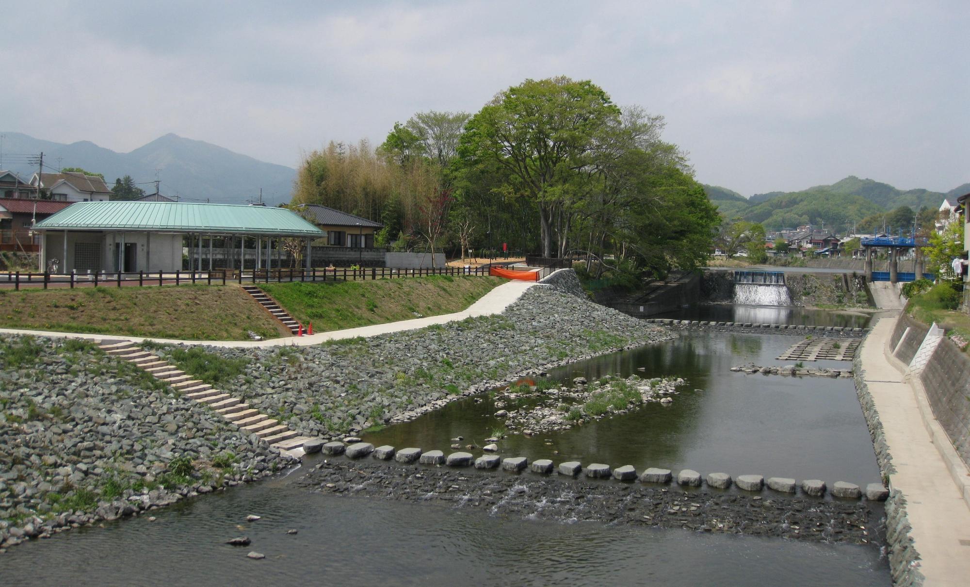 飛び石が設置された川の左側に薄い緑色の大きな屋根の東屋やトイレが完備されている栃本親水公園の写真