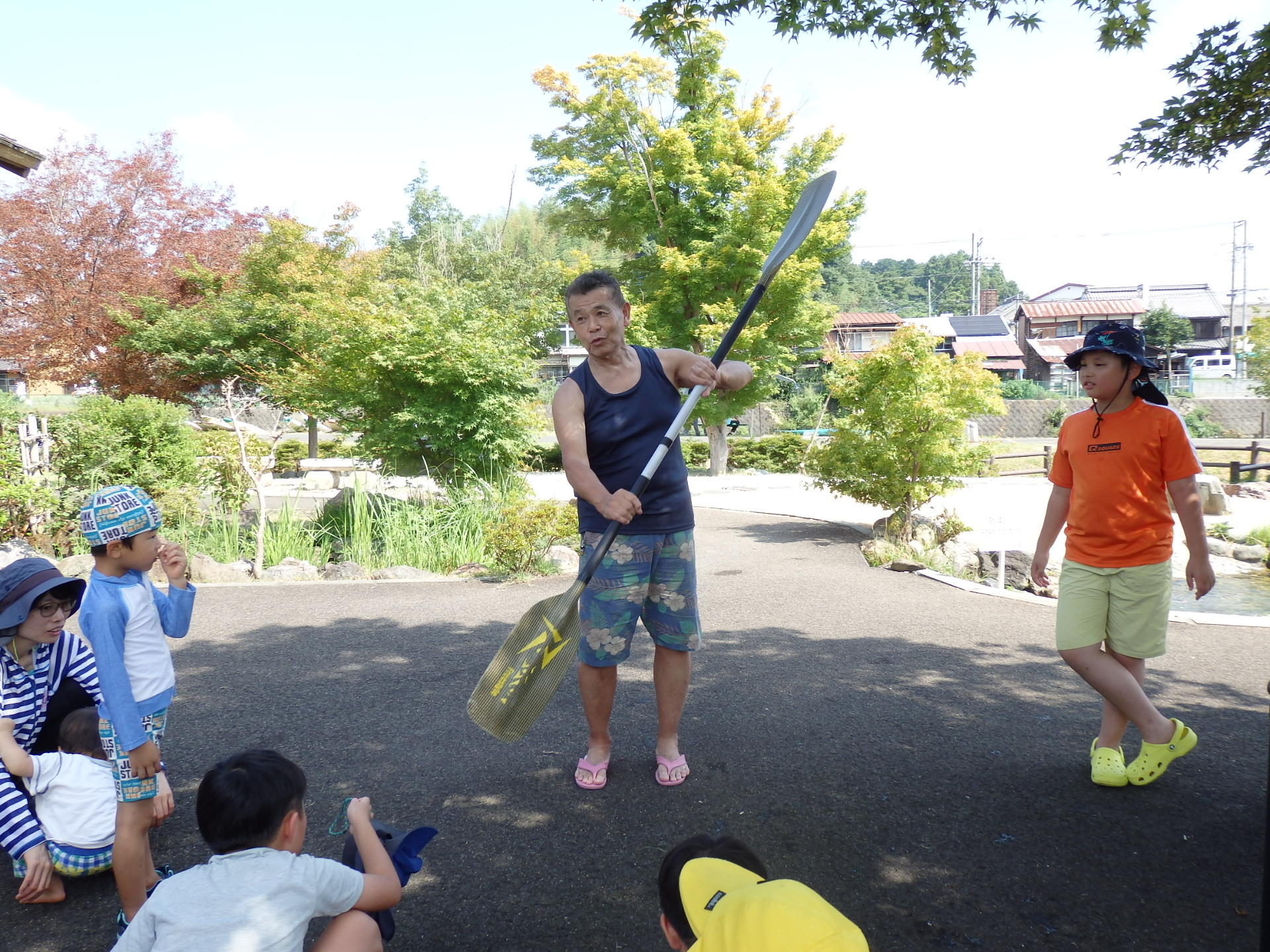 木陰の下に集まった参加者達が、両手でパドルを持った男性の説明を聞いている写真