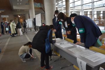 長机に用紙がセットされていて3名の関係者が立ち向かい合わせの女性が中腰で用紙を記入している受付の様子を撮影した写真