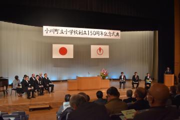 小川町立小学校創立150周年記念式典と書かれた横断幕が掲げられたステージの上で壇上の両側に座る関係者の様子を撮影した写真
