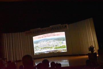 照明が落とされたステージ上でスクリーンに映し出された映像を後方から撮影した写真