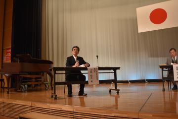 ステージ上のテーブルについた男性が手にマイクを持っている様子を撮影した写真