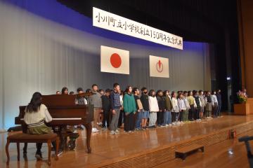 白いシャツを着た女性がピアノを演奏し、ステージ上で合唱する児童たちを左方向から撮影した写真