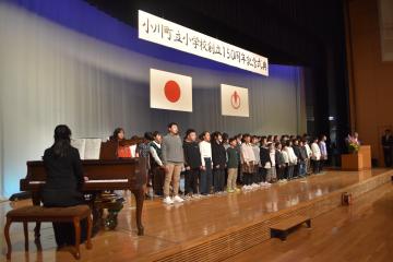 黒い服を着た女性がピアノを演奏し、ステージ上で合唱する児童たちを左方向から撮影した写真