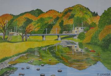 山の木々の葉がオレンジに色づいた様子や麓に立つ建物と橋が、川の水面に反射している様子を描いた内田 敏雄さんの作品