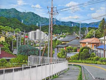 緩やかな傾斜の道の先に広がる家々と山並みの風景が描かれた長谷部 紀一郎さんの水彩画の写真