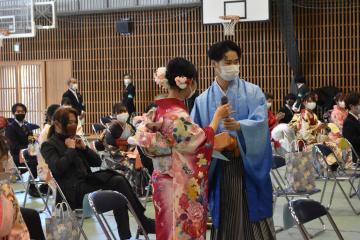 パイプ椅子の前で振袖姿の女性と羽織袴姿の男性がマイクをいっしょに持ち立っている写真