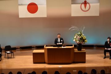 日本国旗と小川町の町章旗を背景に町長が演台の置かれたスタンドマイクの前で式辞を述べている様子の写真