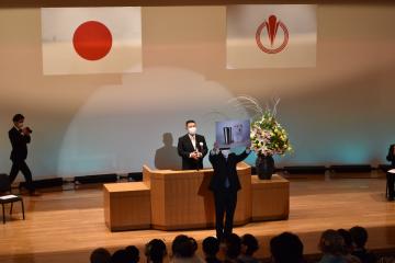 演台の前に立つスーツ姿の男性の前方で記念品を両手に持ち上に掲げて参加者に見せているスーツ姿の男性の写真