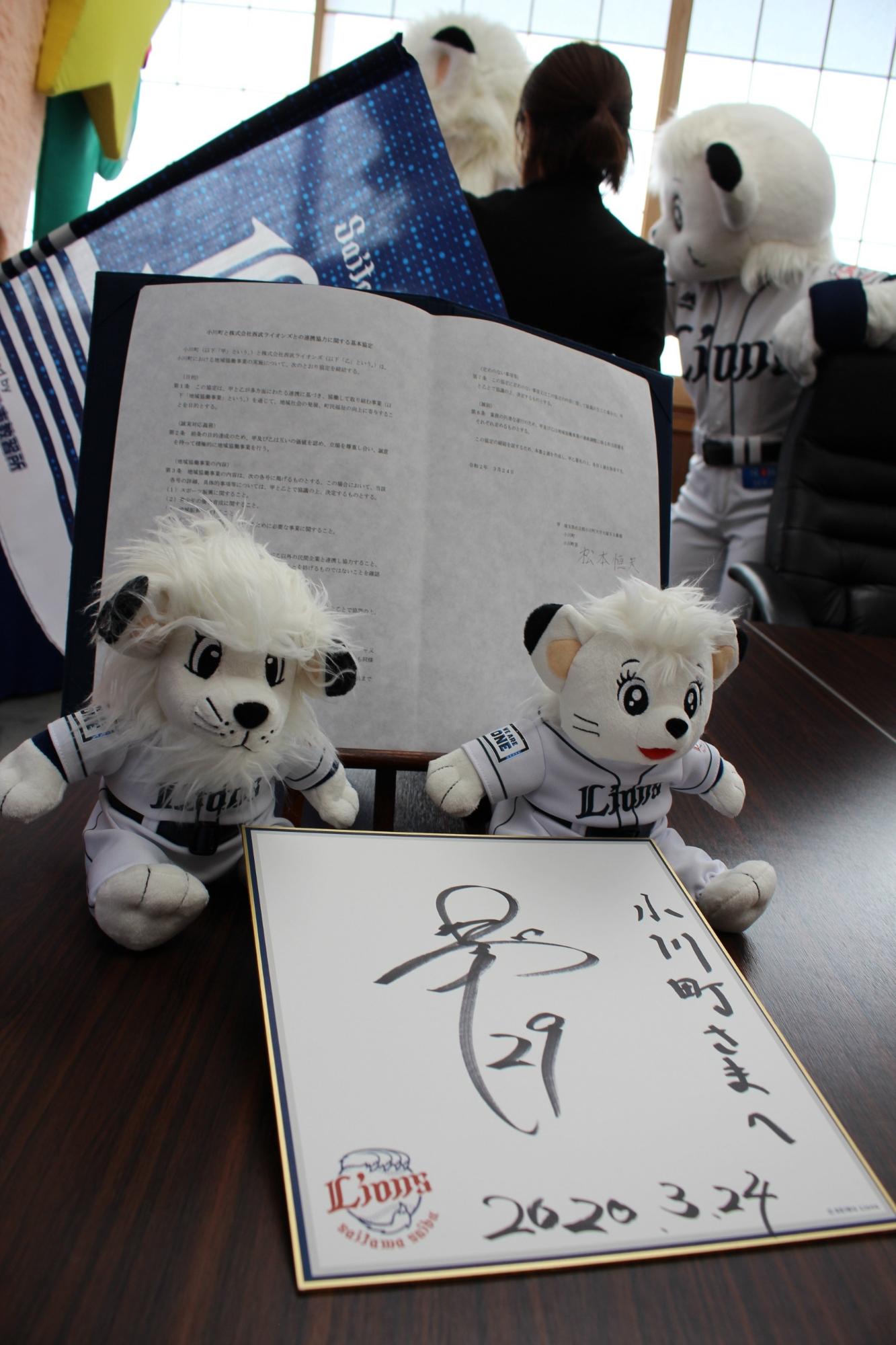テーブルに置かれた協定書、球団マスコットのレオとライナのぬいぐるみそして小川投手のサイン色紙の写真