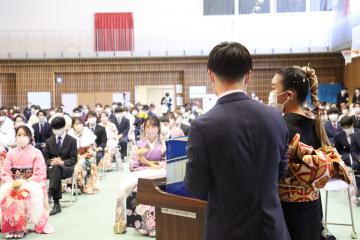 参加者が着席している会場内の一角に置かれた演台の前でスーツ姿と振袖姿の男女が立っている写真