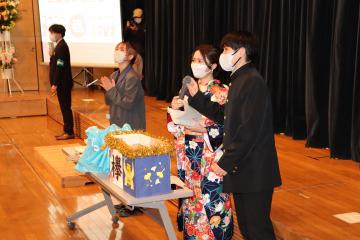 長テーブルの上に景品が置かれておりそのテーブルの前で学生服を着た関係者がマイクを持ち振袖姿の女性がそのマイクに向かって呼びかけている様子の写真