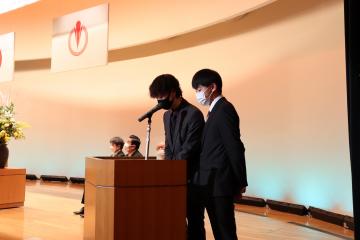 ステージの端に置かれた演台の前で2名のスーツ姿の男性が話をしている様子の写真