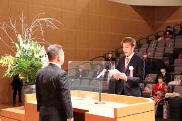 演台をはさみ向かい合わせ立っている町長と式辞表を持ち誓いのことばを述べているスーツ姿の男性の写真