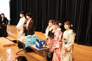 長テーブルの上に景品が置かれており羽織袴や振袖姿の関係者が立っている様子の写真