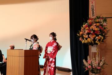 花輪が置かれたステージ上の隣で2名の振り袖姿の女性が演台の前に立っている様子の写真