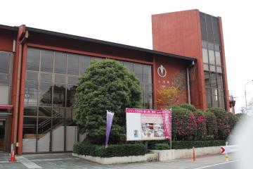 茶色の外壁で全面ガラス張りになっている建物で建物に沿って植樹された花壇などがあるリリックおがわ外観写真