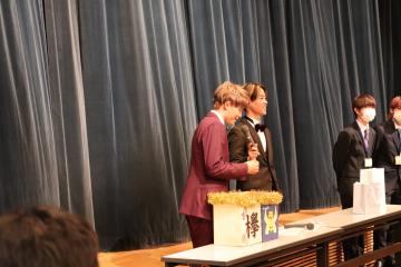 長テーブルの上に装飾された箱が置かれておりスーツ姿の関係者2名が立っている写真