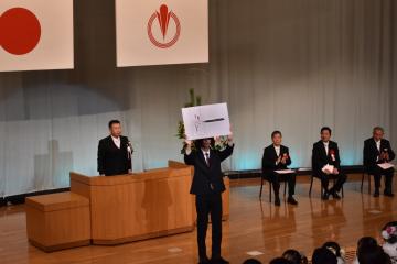 演台の前に立つスーツ姿の男性の前方で記念品を両手に持ち上に掲げて参加者に見せているスーツ姿の男性の写真